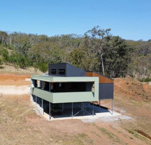 Modern elevated house on stilts in a cleared hillside, with a green exterior and dark upper section, surrounded by trees.