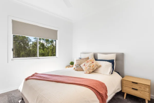 Bright bedroom with a neatly made bed, cream bedding, peach throw, decorative pillows, two wooden nightstands, and a window showing trees outside.