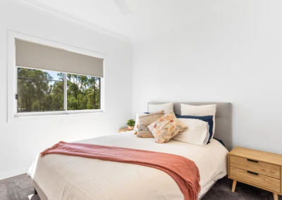 Bright bedroom with a neatly made bed, cream bedding, peach throw, decorative pillows, two wooden nightstands, and a window showing trees outside.
